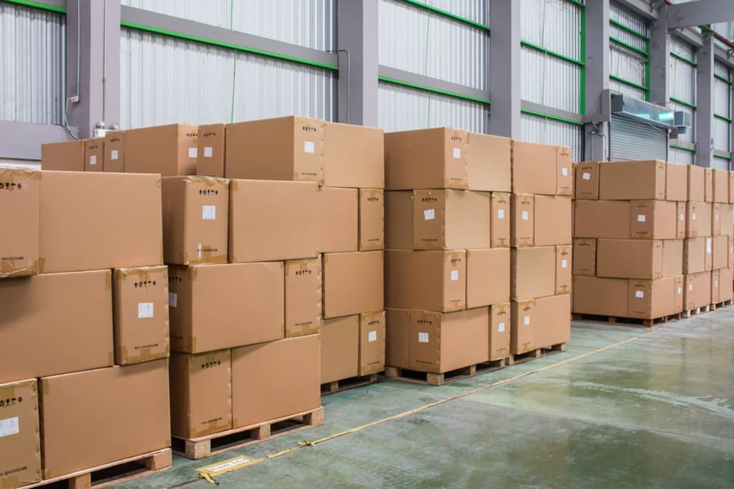 Stocktake Sydney Asset Valuations, Stacks of large cardboard boxes on wooden pallets are arranged in rows inside a warehouse with high ceilings, metal walls, and concrete floors during a Stocktake Sydney inventory check.