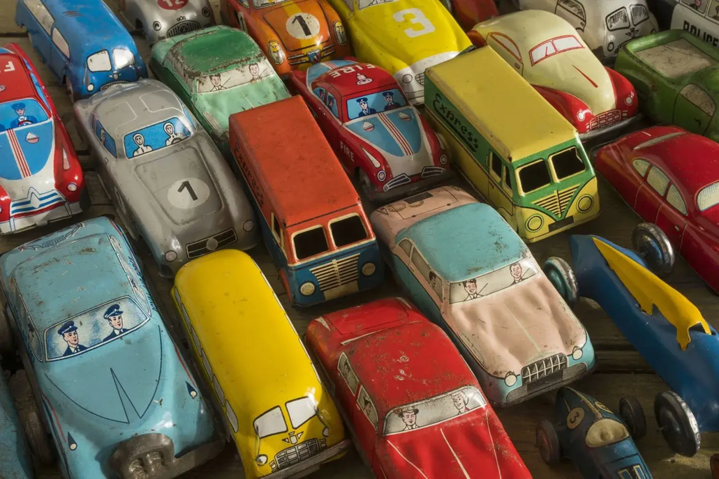 A collection of colorful vintage toy cars, buses, and vans made of metal are arranged closely together, showing signs of wear recognized in Toy Grading Terminology Australia. Some vehicles feature painted drivers and numbers on their roofs.