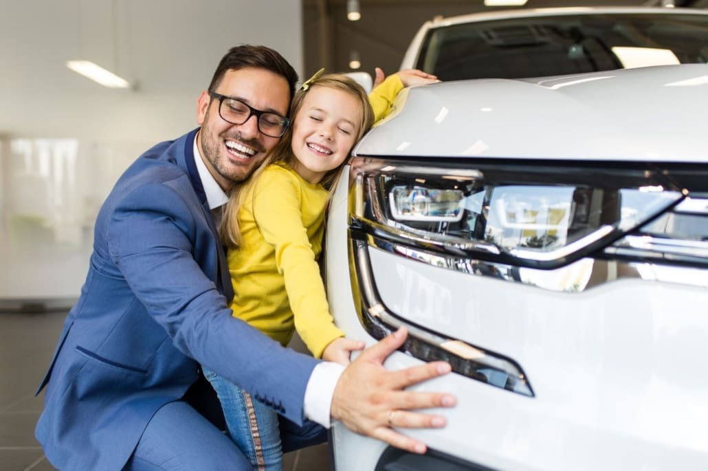 A man in a blue suit and a young girl in a yellow sweater smile happily while hugging the front of a white car inside a VALUATIONS BRISBANE showroom.