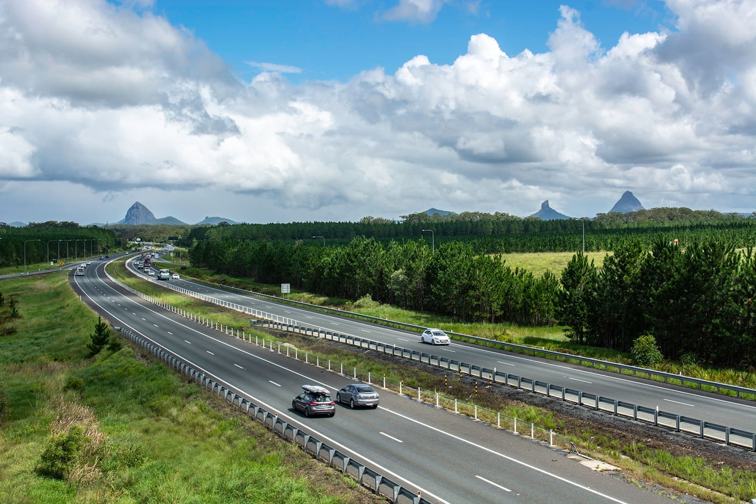 A divided highway with cars traveling in both directions near Valuations Rockhampton, surrounded by green trees and fields under a partly cloudy sky, with distant mountains visible on the horizon.