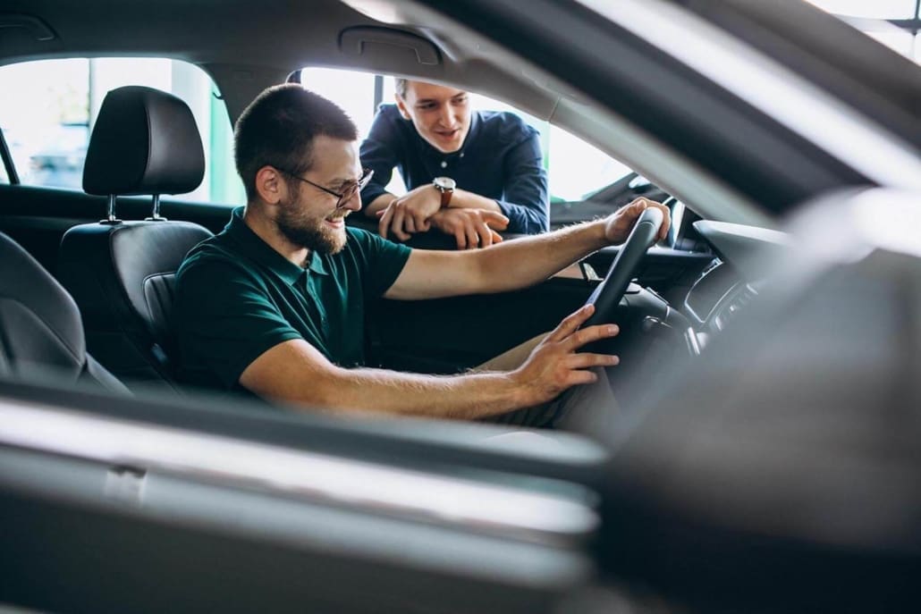 A man sits in the driver’s seat of a car, smiling and holding the steering wheel, while another man leans in from outside the car window discussing vehicle valuations Brisbane inside a showroom.