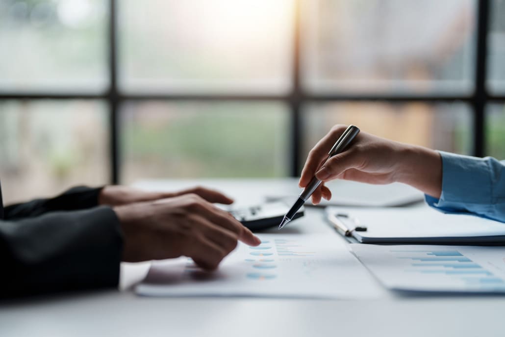 Two people’s hands discussing data at a table with printed charts and graphs, one person pointing with a pen. A clipboard and documents—including info on What Are Asset Registers—are visible, with large windows in the background.