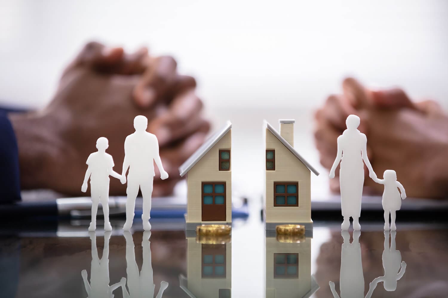 Family-Law-Valuations-AVG Two people sit across from each other with hands clasped, behind paper cutouts of a divided house and separated family figures, symbolizing separation or divorce and family division.
