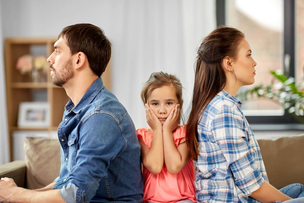 When Are Family Law Valuations Required When Are Family Law Valuations Required - A man and woman sit back-to-back on a couch, looking away from each other. A young girl sits between them, holding her face in her hands and looking concerned. The scene suggests family tension.