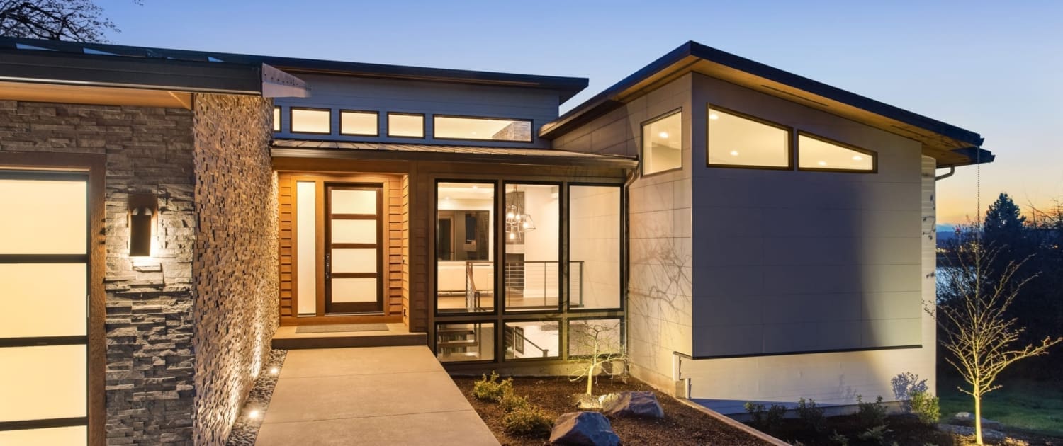 Property Valuations Broome, Modern house exterior at dusk with large windows, stone and wood accents, illuminated walkway, landscaped front yard, and a stunning sky in the background—ideal for those seeking Property Valuations Broome.