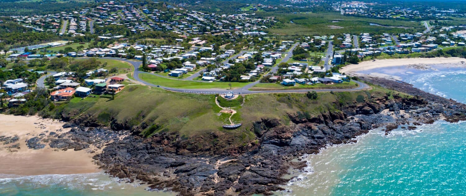 Aerial view of a coastal town with houses near green hills, rocky cliffs, and sandy beaches—perfect for Property Valuations Bundaberg—bordered by turquoise ocean waves under a blue sky with scattered clouds.