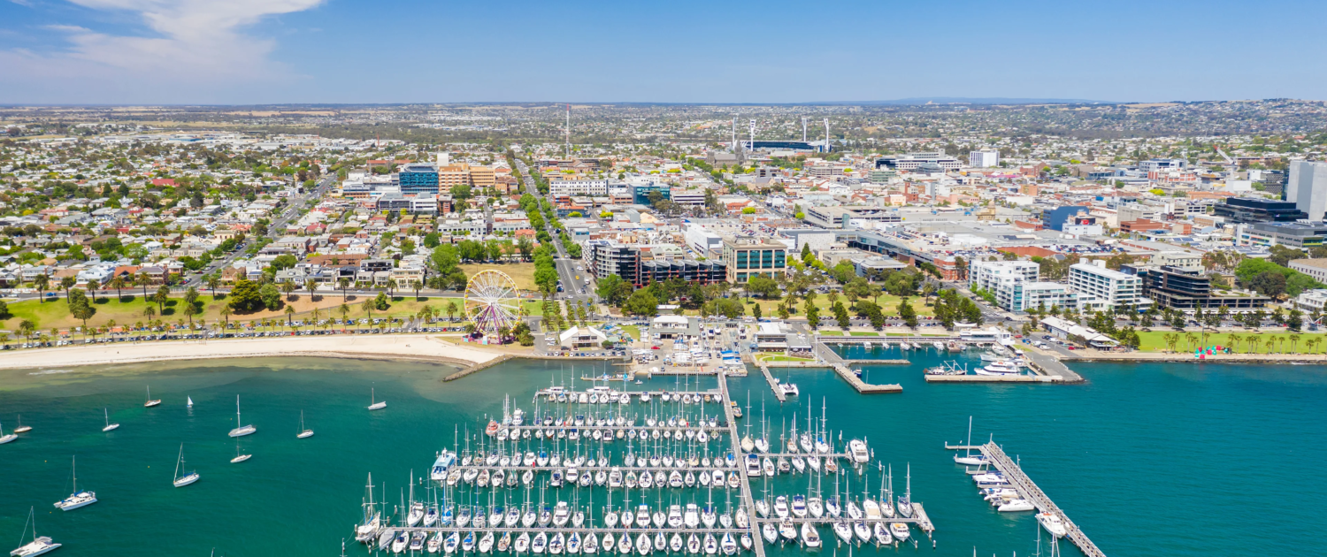 Property Valuations Geelong, Aerial view of a marina filled with boats and yachts on blue water, with a sandy beach, green park, and the Geelong cityscape in the background—perfect for those interested in Property Valuations Geelong under a clear blue sky.