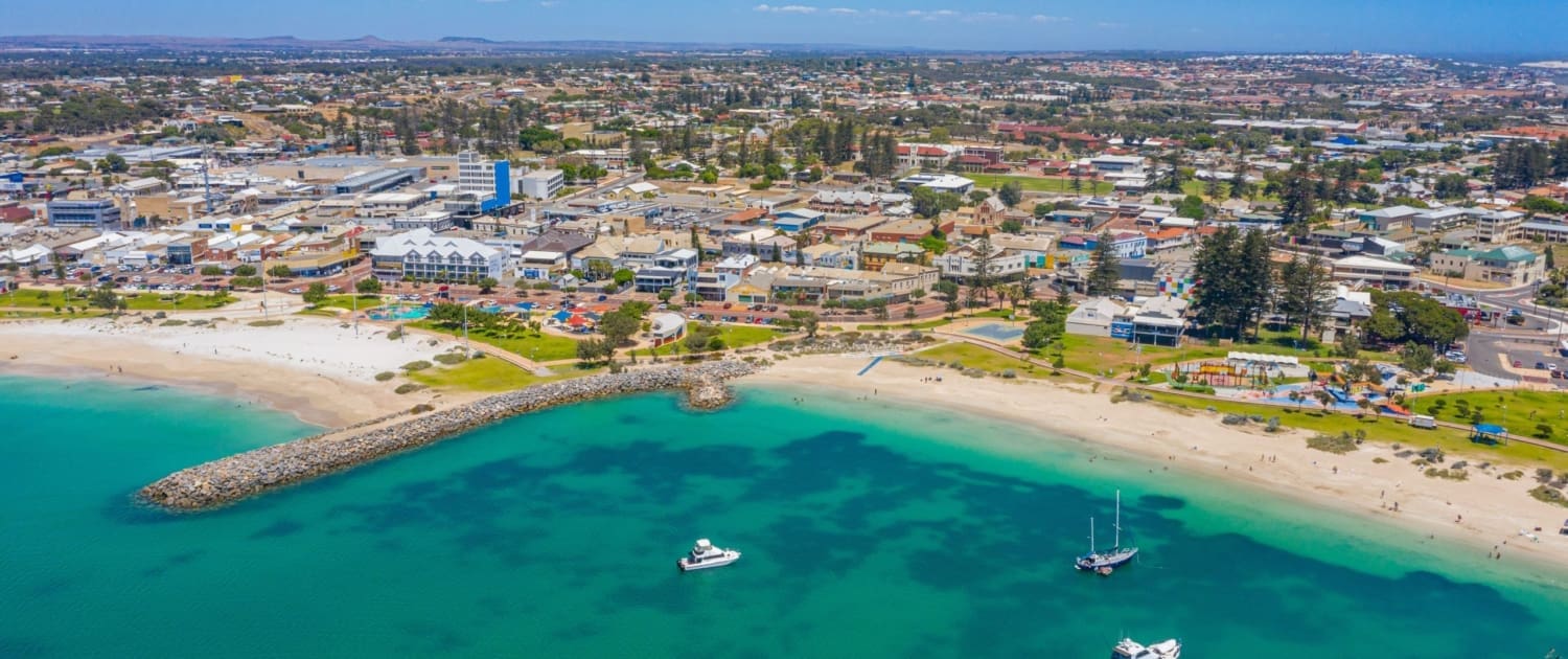 Property Valuations Geraldton, Aerial view of a coastal town with sandy beaches, turquoise water, a jetty, and boats anchored near shore. Buildings and streets stretch inland under a clear blue sky—perfect for those considering Property Valuations Geraldton.