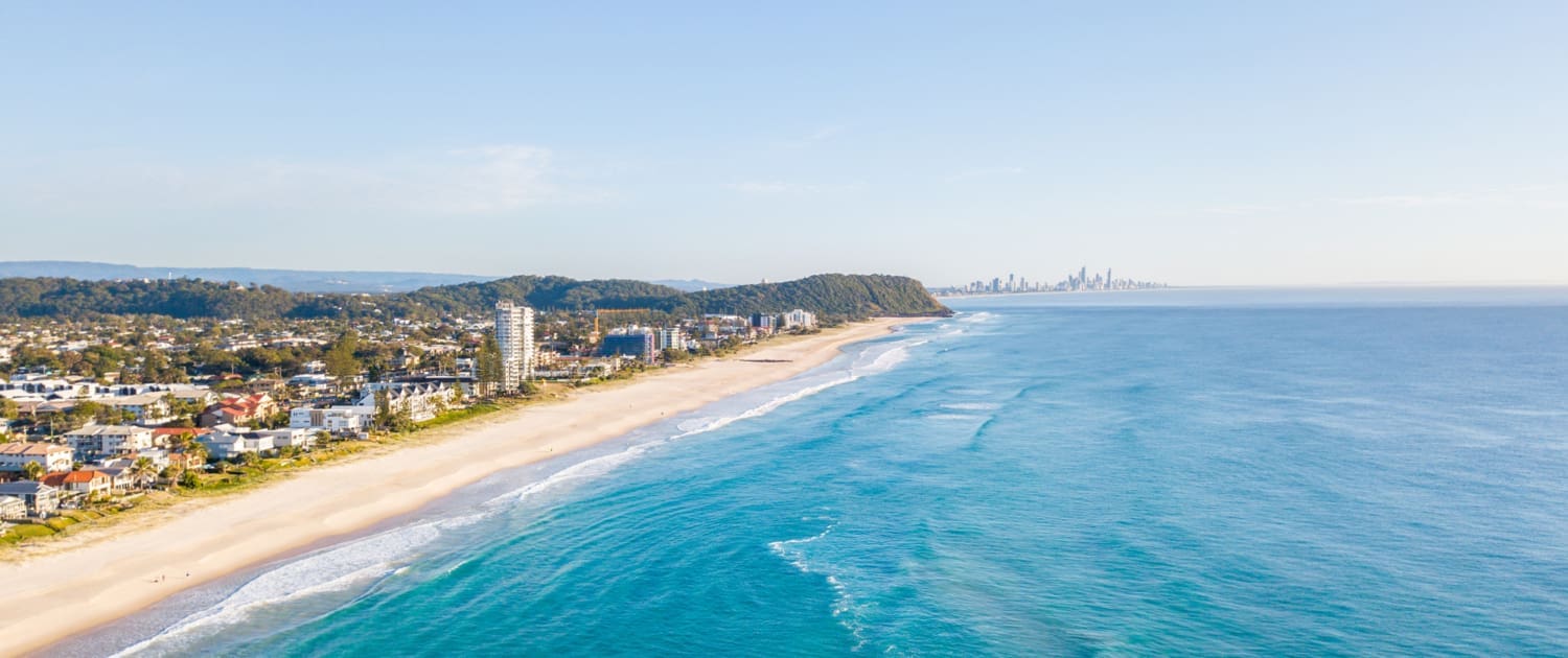 Property Valuations Gold Coast, Aerial view of a long sandy beach with turquoise waves, buildings and greenery along the shore, and a distant city skyline under a clear blue sky—an ideal setting for Property Valuations Gold Coast.
