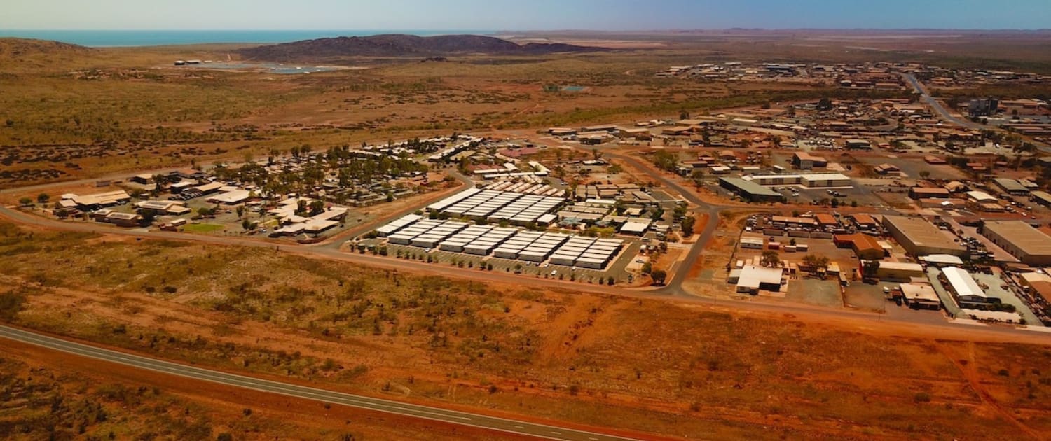 Property Valuations Karratha, Aerial view of a small town surrounded by red earth and sparse vegetation, with grid-like buildings, a highway in the foreground, and distant hills under a clear blue sky—ideal for Property Valuations Karratha.