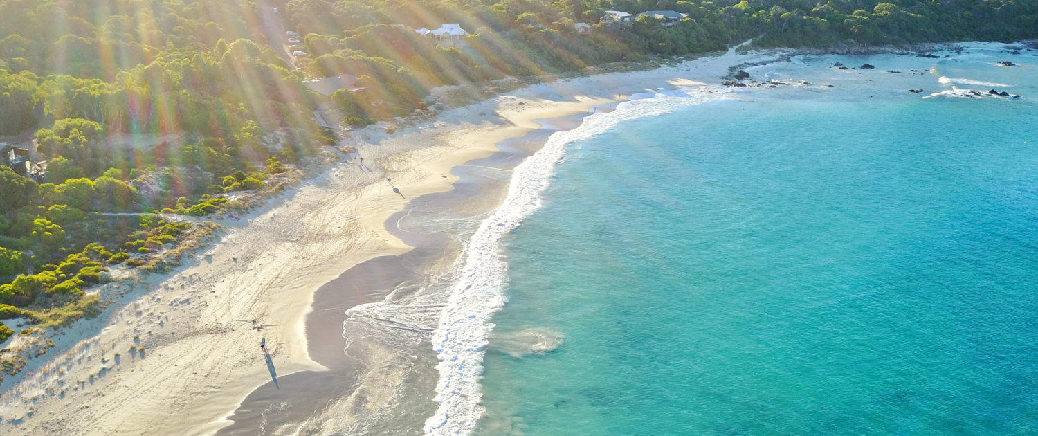 Property Valuations Margaret River, Aerial view of a sunlit beach with gentle waves, light sand, and lush green forest bordering the shoreline. Sun rays stream from the upper left, highlighting this tranquil coastal scene—ideal for Property Valuations Margaret River.