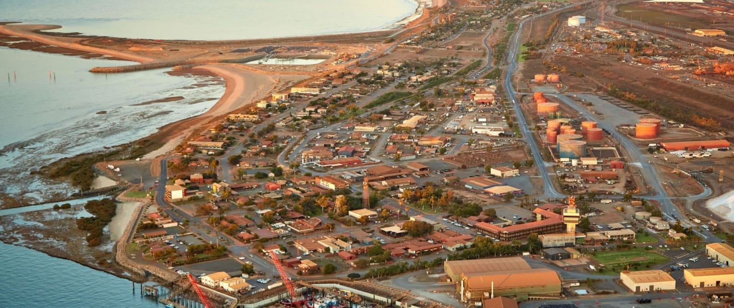 Property Valuations Port Hedland Property Valuations Port Hedland, Aerial view of a coastal town with a marina, boats docked at piers, sandy shoreline, scattered buildings, and industrial tanks—ideal for Property Valuations Port Hedland—with expansive land and water stretching into the background.