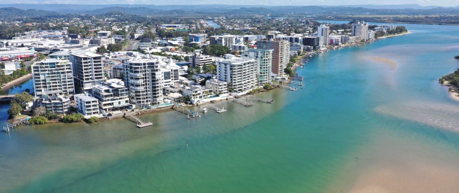 Property Valuations Sunshine Coast, Aerial view of waterfront apartment buildings along a curving river on the Sunshine Coast, with clear blue-green water, sandy shorelines, and a cityscape in the background—ideal for those interested in Property Valuations Sunshine Coast.