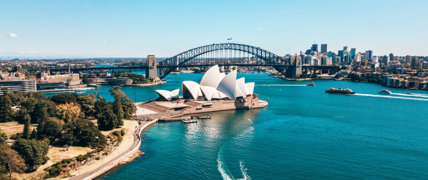 Property Valuations Sydney, Aerial view of Sydney Harbour featuring the Sydney Opera House, Harbour Bridge, and city skyline—an iconic setting often referenced in Property Valuations Sydney, with boats gliding through vibrant blue water under a clear, sunny sky.