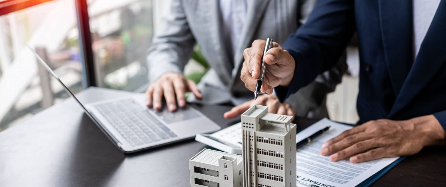 Property Valuers Brisbane, Two people in business attire sit at a desk with a laptop, document, and pen, discussing real estate; a small model of a building is on the table, suggesting Property Valuers Brisbane in action for property or construction planning.