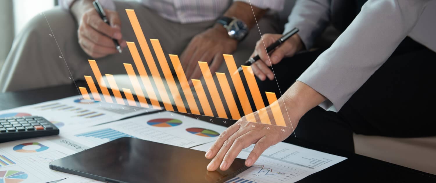 Refinance Valuation Brisbane, Two people review business charts and graphs at a table, with a digital orange bar graph projected above a tablet. Papers with colorful charts, a calculator, and pens suggest detailed financial planning or refinance valuation in Brisbane.