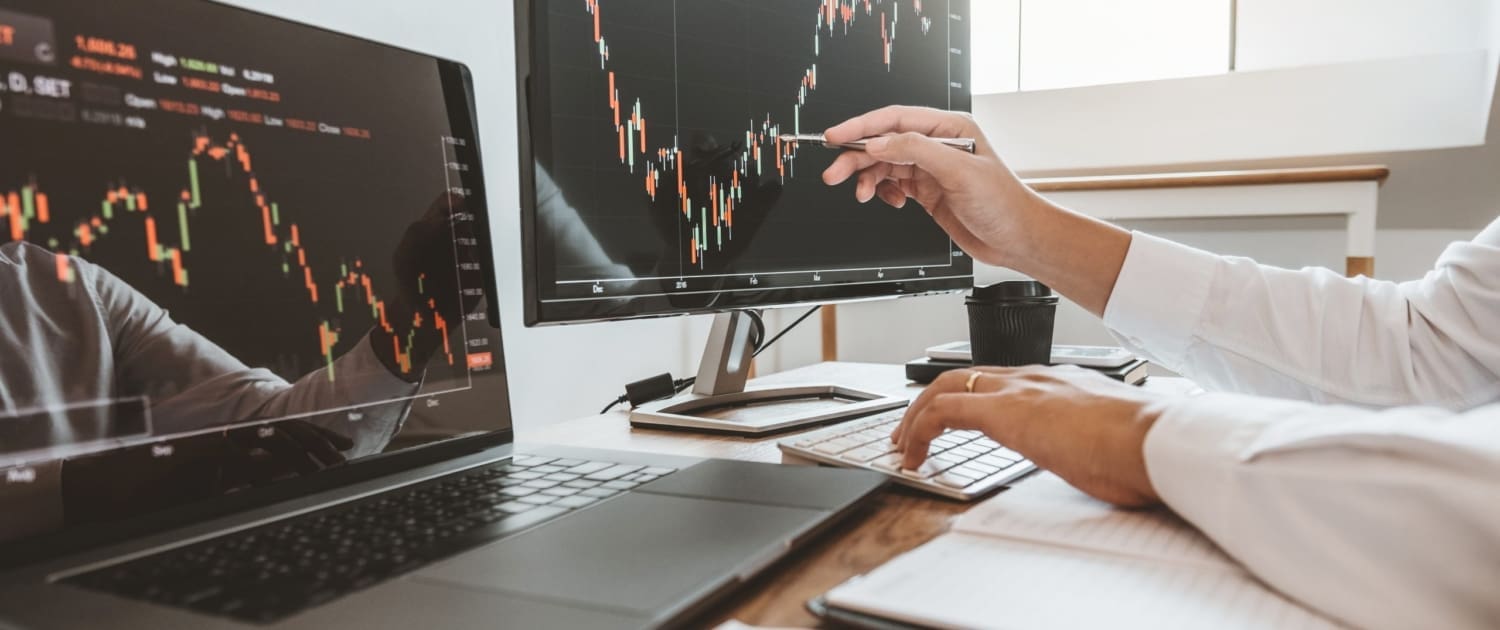 Share Valuations Sydney, Two people analyze share valuations Sydney on financial charts displayed on monitors and a laptop at a desk, using a pen to point at a candlestick graph, with a notebook and coffee cup nearby.