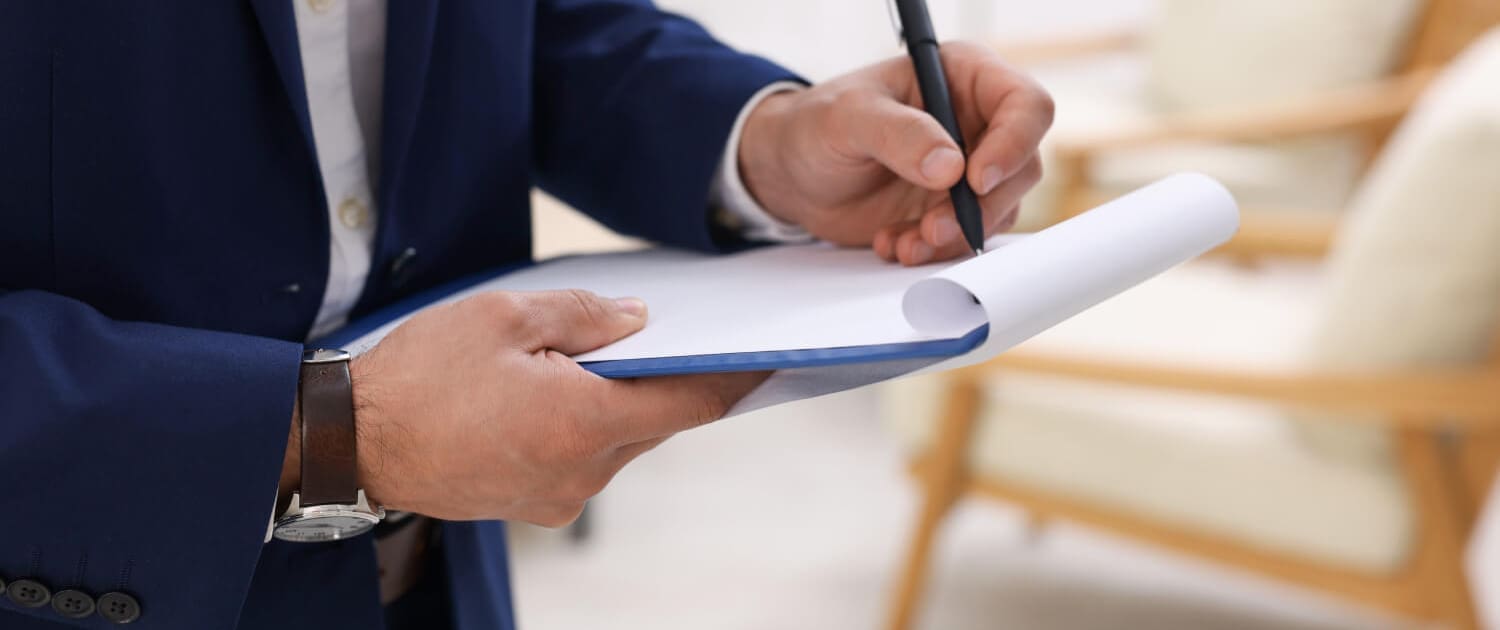 what are asset registers Australia what are asset registers Australia, A person in a navy blue suit holding a clipboard and writing on a sheet of paper, possibly compiling data for an asset register, with a blurred background showing light-colored chairs.