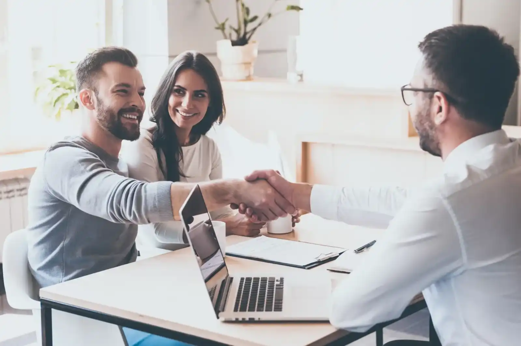 How the Brisbane Property Valuation Process Works, A man and woman sit at a desk, smiling, while the man shakes hands with another man across from them—perhaps celebrating successful property valuations in Brisbane. A laptop, documents, a cup, and a plant complete the inviting setting.