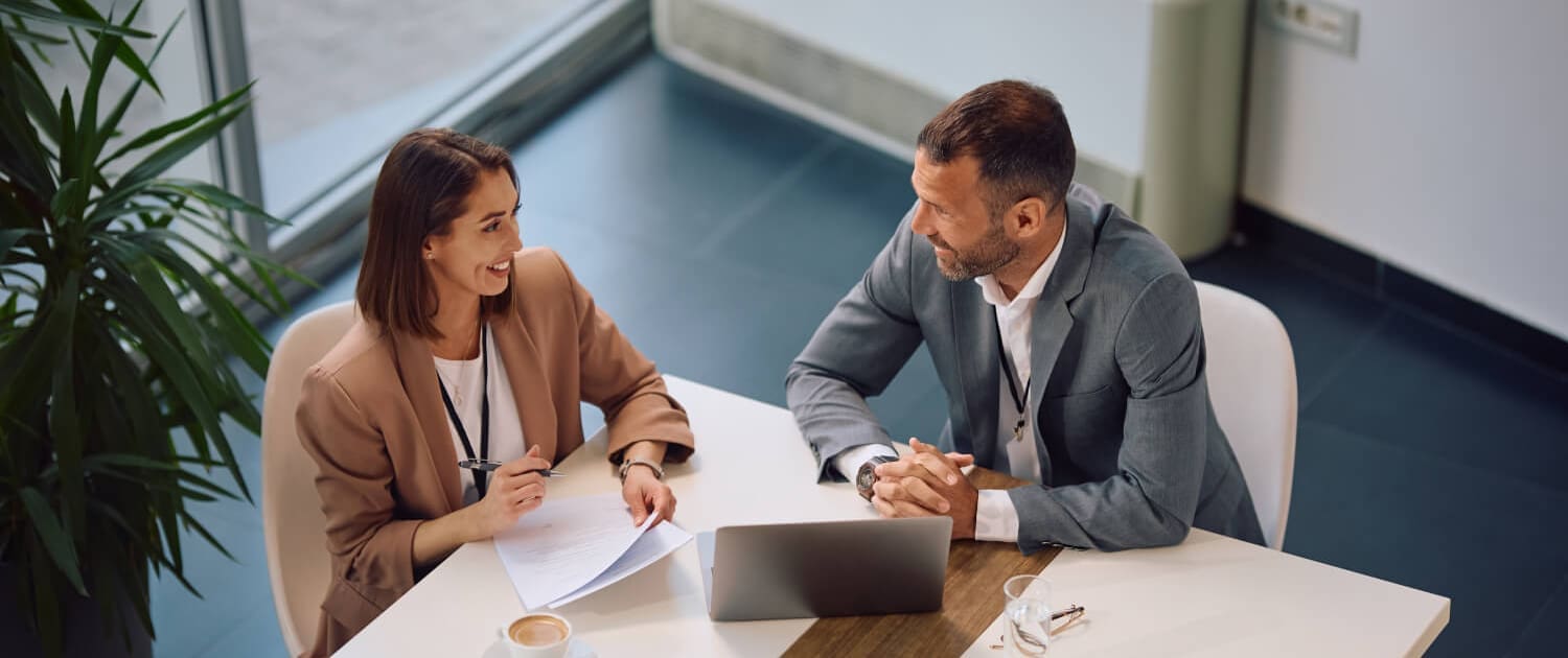 Two business professionals, a woman and a man, sit across from each other at a table in a bright Asset Management Brisbane office, talking and smiling. A laptop, documents, smartphone, coffee, and glasses are on the table.