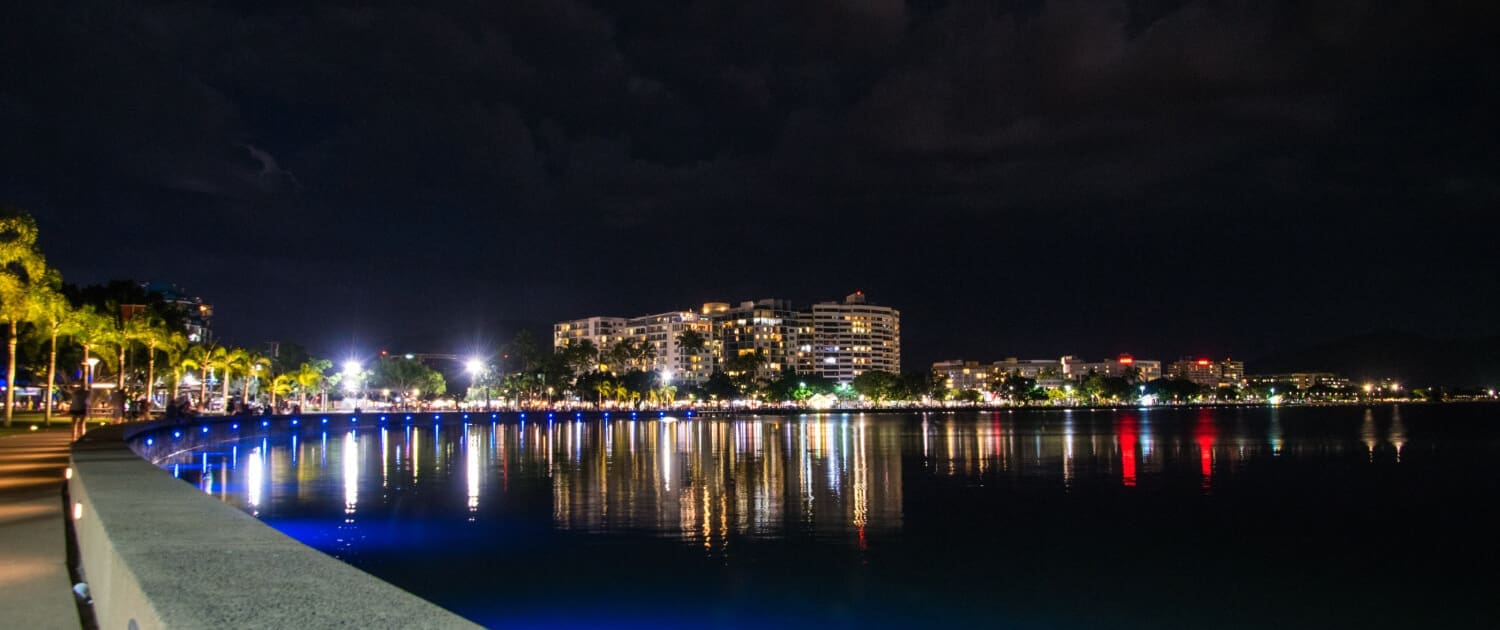 A nighttime view of a waterfront promenade in Cairns with city buildings illuminated by colorful lights—ideal for those seeking Business Valuations Cairns—reflections shimmer on calm water under a dark, cloudy sky.
