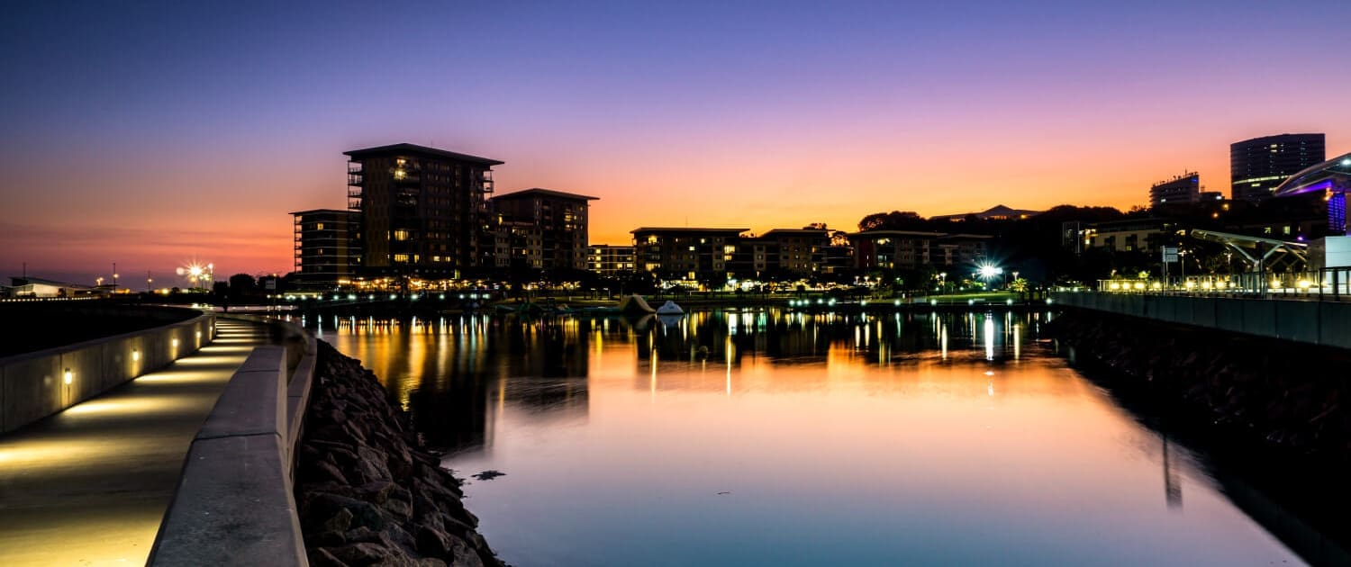 Modern waterfront buildings reflect on calm water at sunset, under a vibrant sky transitioning from orange to deep blue. Along the lit walkway bordered by rocks, the urban scene hints at the thriving growth fueled by Business Valuations Darwin.