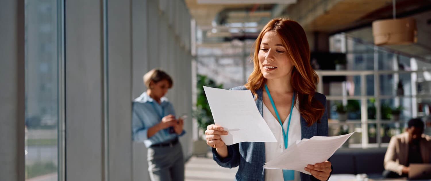 A woman in a blue blazer stands in a modern office, reading documents related to Business Valuations Gympie. Behind her, another person is near large windows and a third sits at a table. The space is bright with natural light.