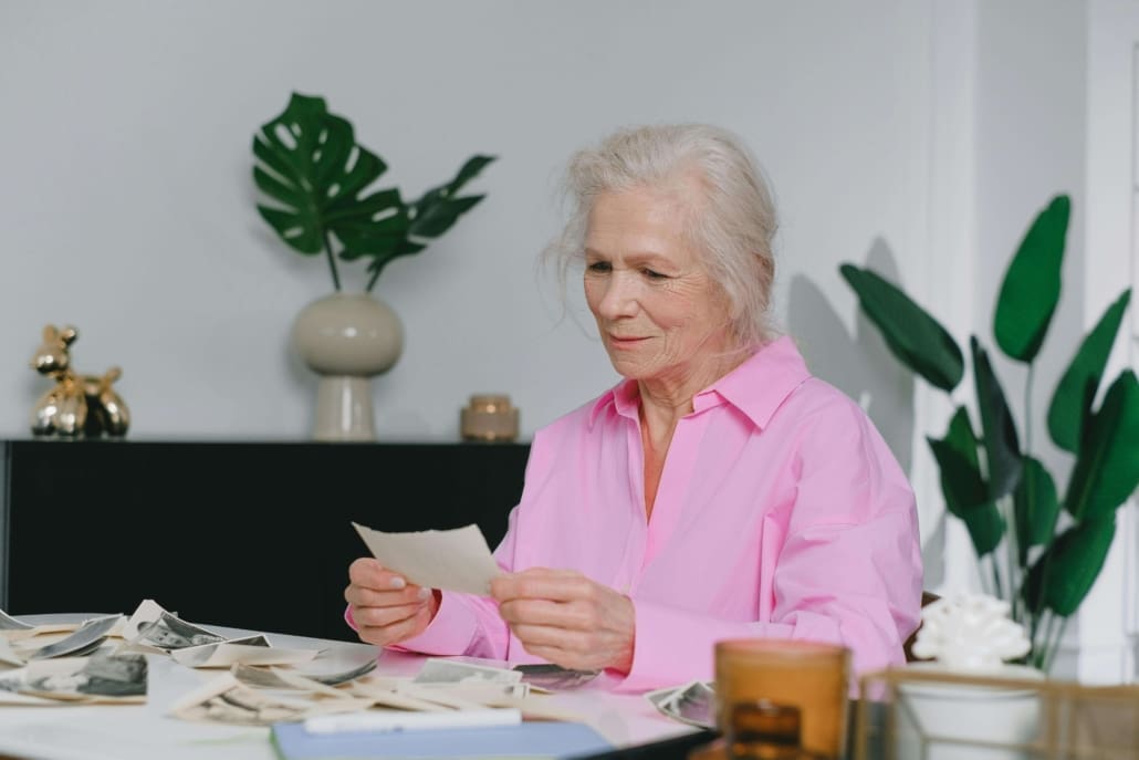 Assets Commonly Requiring Deceased Estate CGT Valuations An elderly woman with gray hair wearing a pink shirt sits at a table, looking at old photographs related to Deceased Estate CGT Valuations. The table is scattered with more photos, and green plants are in the background.