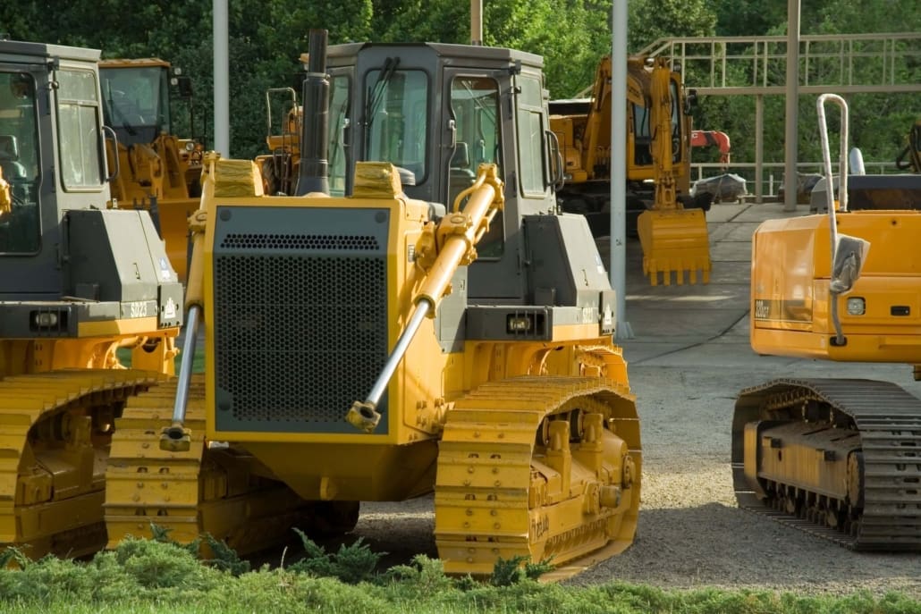 Several yellow bulldozers and construction vehicles are parked outdoors on gravel near green bushes and trees, with one bulldozer facing the camera in the foreground—ideal for Plant & Equipment CGT Valuations.
