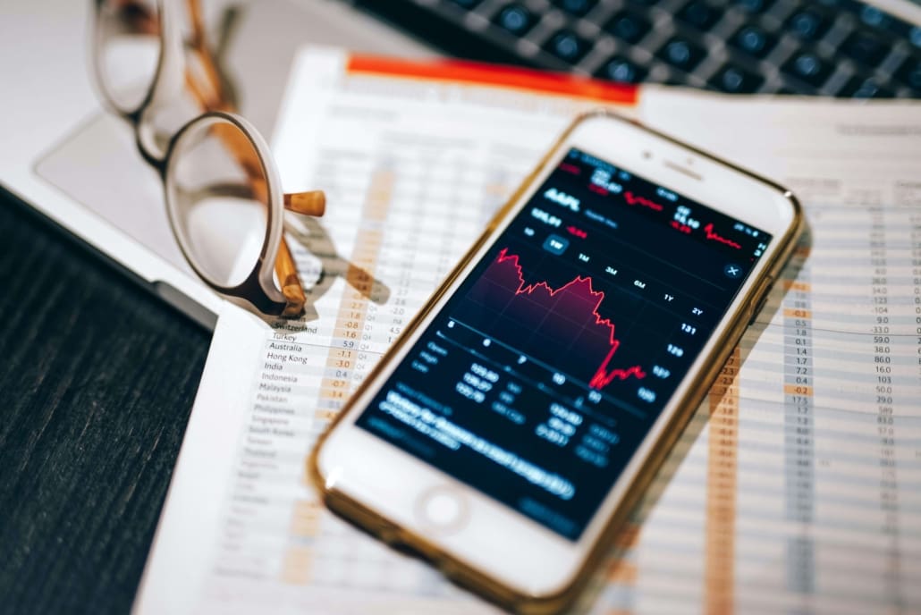 A smartphone displaying a stock market chart lies on financial documents next to eyeglasses and a laptop keyboard, highlighting fluctuating data in red—ideal for monitoring shares & equity CGT valuations during potential declines.