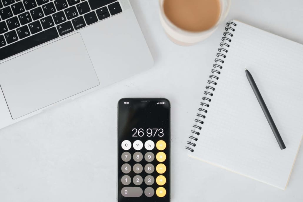 CGT-Valuations-Australia A smartphone displaying the number 26,973 on a calculator app lies on a white desk next to a laptop, spiral notebook with pen, and coffee cup—tools often used for Capital Gains Tax Valuations.