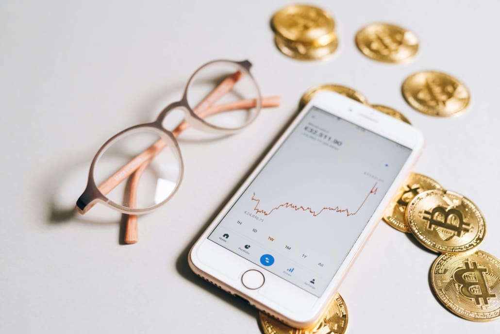 Intangible-Asset-CGT-Valuations A smartphone displaying a rising cryptocurrency chart, surrounded by physical Bitcoin coins and a pair of eyeglasses on a light surface, illustrating concepts like Goodwill & Intangible Asset CGT Valuations in digital finance.