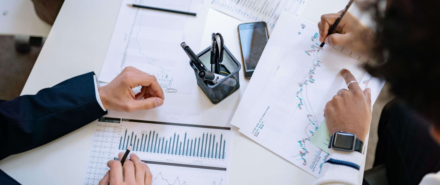 Business Interest CGT valuations Three people sit at a desk reviewing printed charts and graphs, with pens, smartphones, and a laptop on the table, suggesting a business or financial meeting focused on Business Interest CGT Valuations.