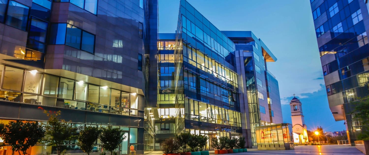 Modern glass office buildings at dusk with illuminated windows surround a spacious courtyard with potted plants; a clocktower is visible in the background under a blue evening sky—ideal for Commercial Building CGT Valuations.
