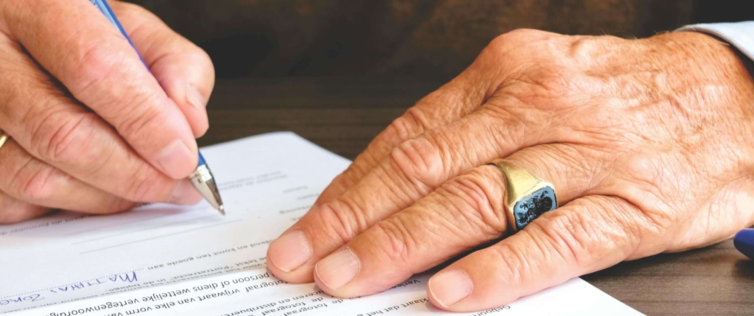 deceased Estate CGT valuations Close-up of an elderly person’s hands signing a document related to Deceased Estate CGT Valuations; one hand steadies the paper, displaying a gold ring with a dark stone, while the other hand writes on the form.