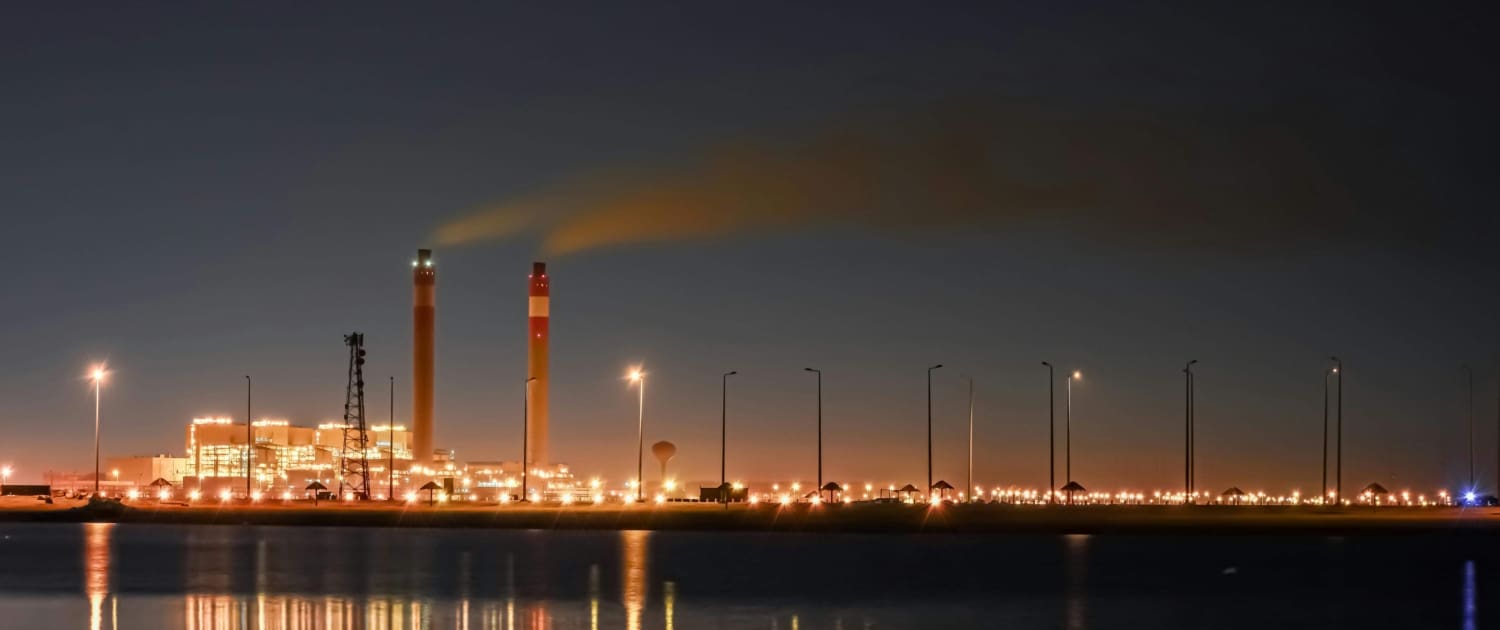 A factory with two tall chimneys emits smoke at night, illuminated by bright lights. The industrial complex, ideal for Industrial Asset CGT Valuations, is reflected in calm water in the foreground under a dark sky.