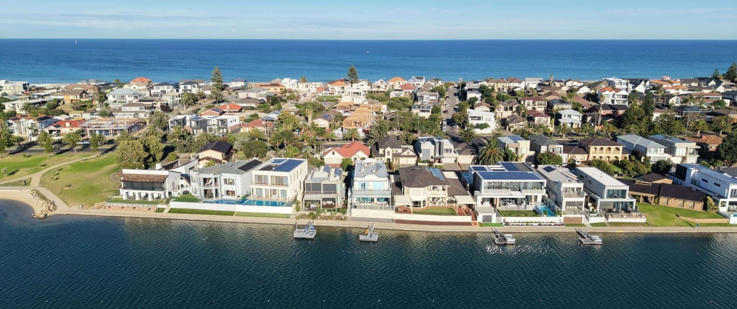 Aerial view of a coastal suburban neighborhood with modern waterfront homes, private docks, and calm water in the foreground—ideal for Residential Investment Property CGT Valuations. The ocean and blue sky create a stunning backdrop.