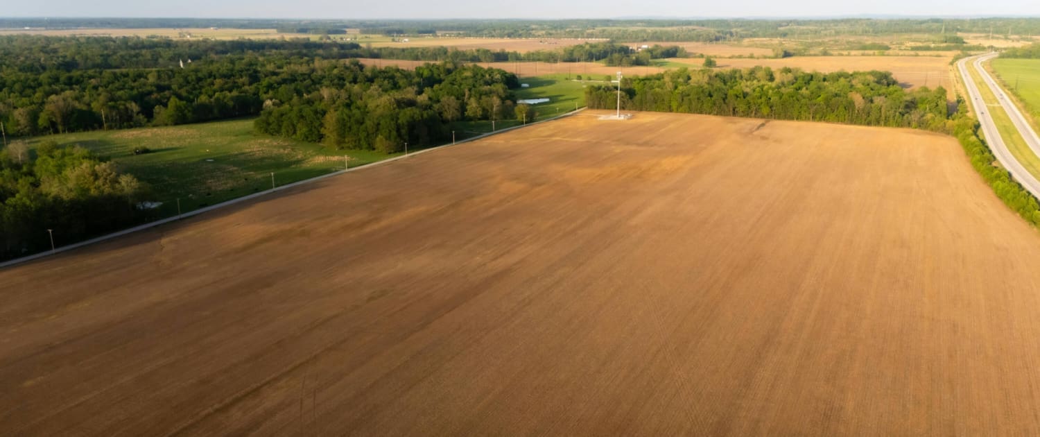 Aerial view of a large, freshly plowed field ideal for Vacant Land CGT Valuations, bordered by green trees and a country road, with distant farmland and a blue sky on the horizon.